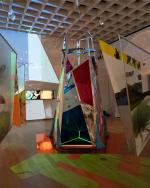 Teepee made of multicolor fabrics in center of wood floored gallery with suspended fabric triangles patterned with geometric elements and green shapes projected across walls and floor.