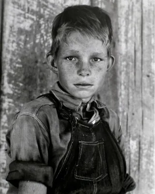 A blond boy in overalls and rolled up sleeves looking directly at the viewer.